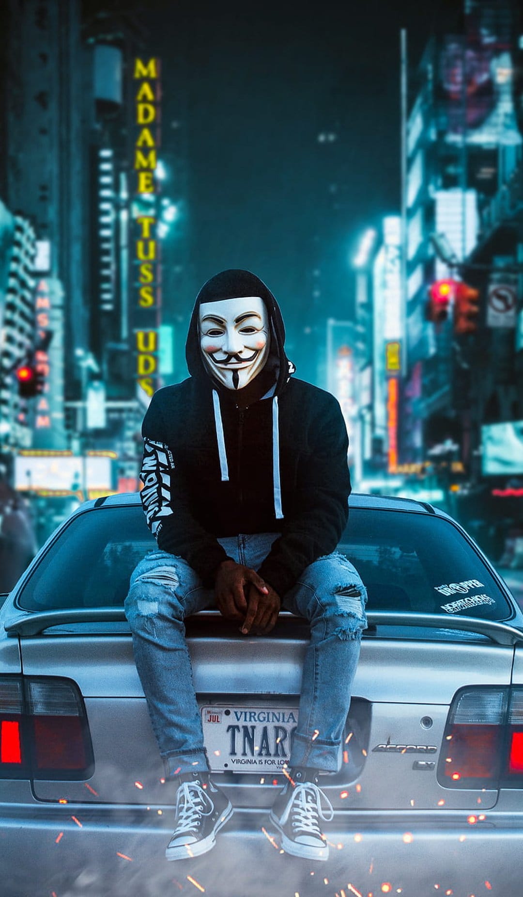 Anonymous masked hacker in a black hoodie sitting on a car trunk against a glowing neon cyberpunk city backdrop.