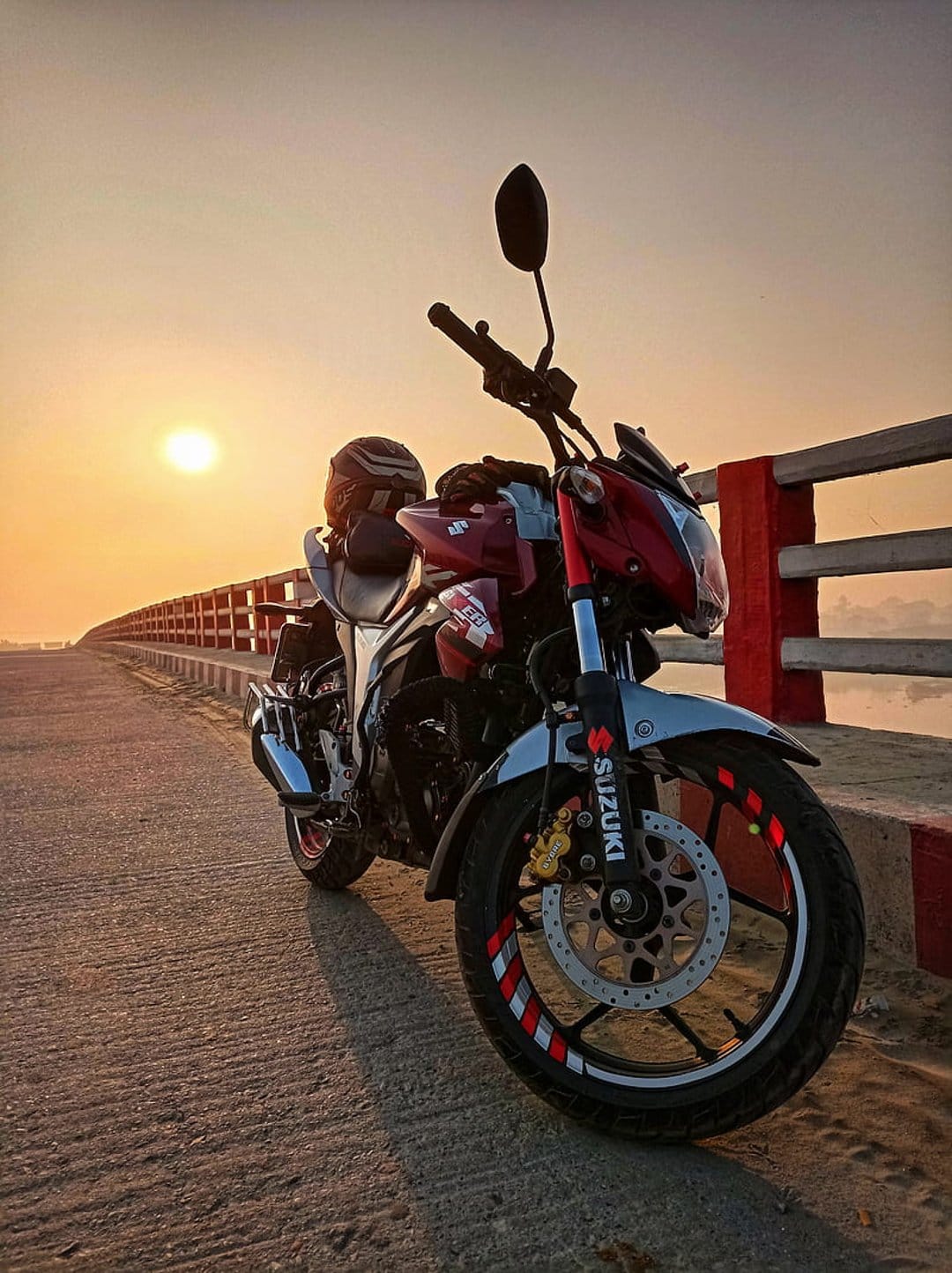 Suzuki motorcycle with red wheel accents sits on a bridge under a warm golden sunset with a soft glowing horizon.