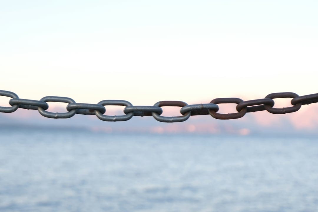 Oxidized metal chain links stretch across a calm blue sea under a soft pink and orange sunset horizon.