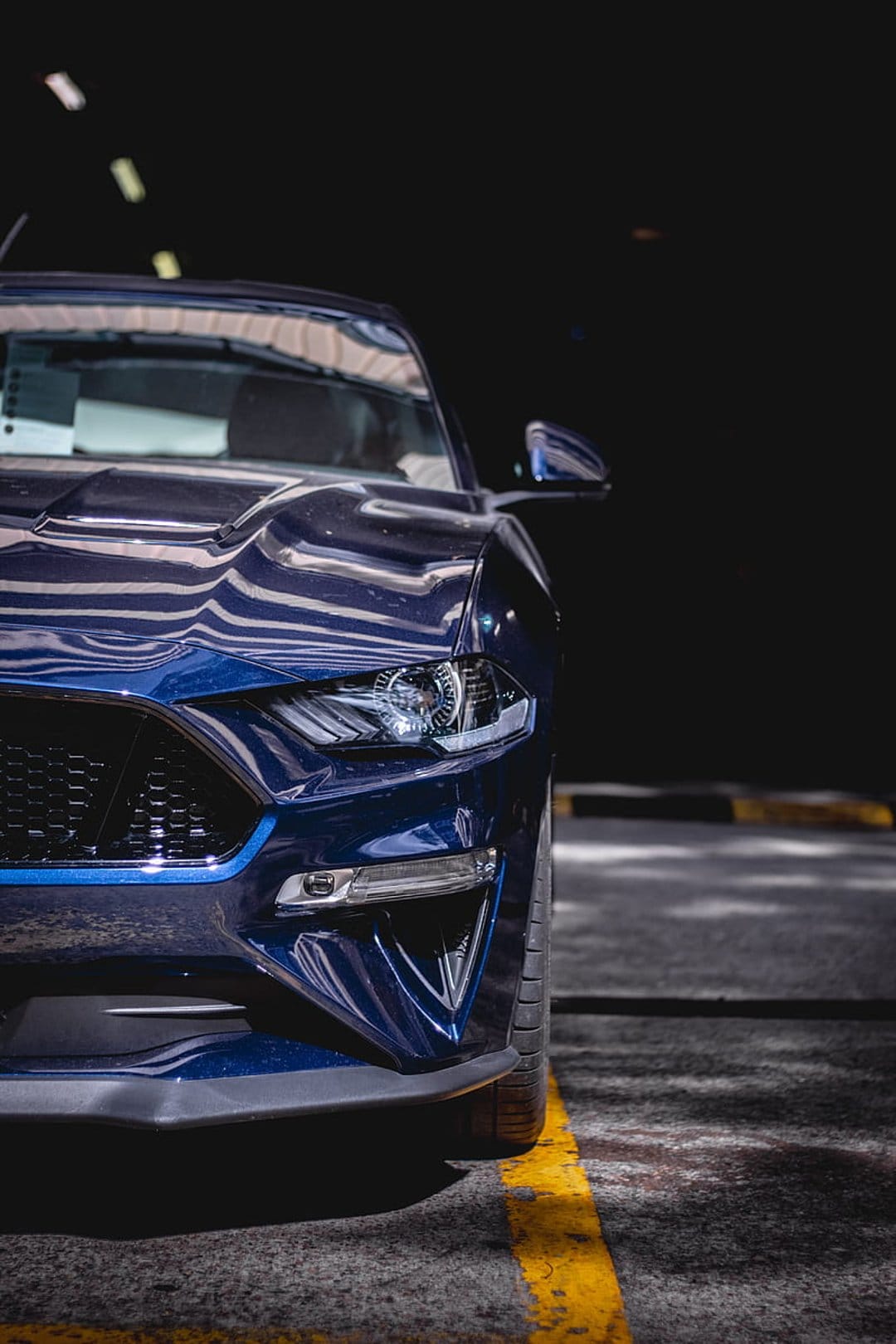 Blue Ford Mustang headlight and grille close-up with glossy reflections and blurred garage background.