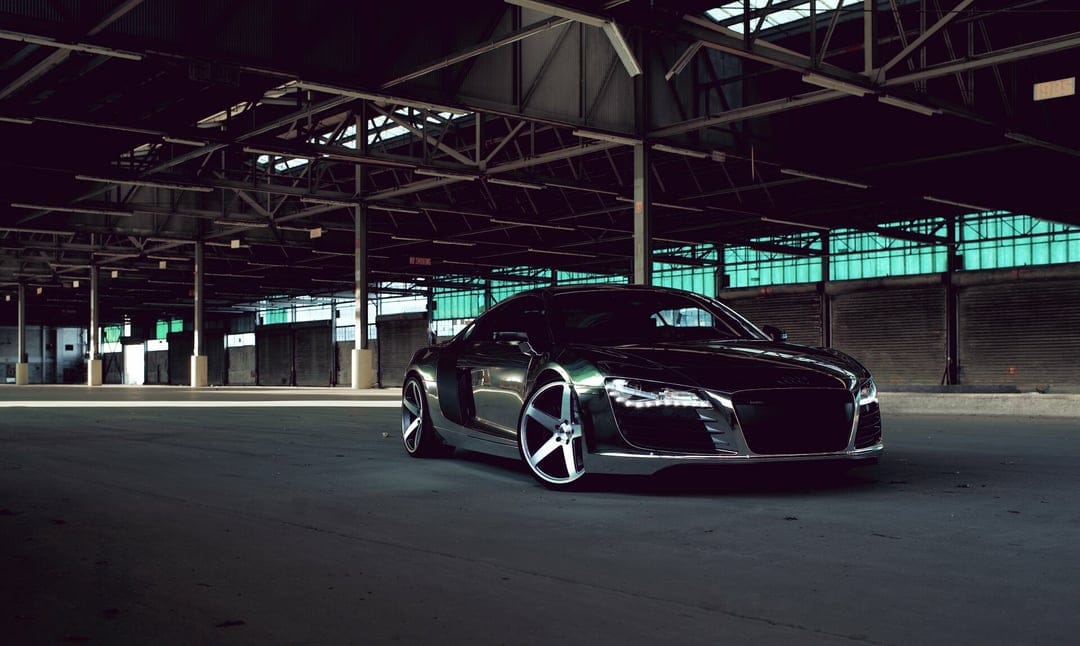 Chrome Audi R8 with glowing headlights parked in a dark warehouse with steel rafters and teal windows.