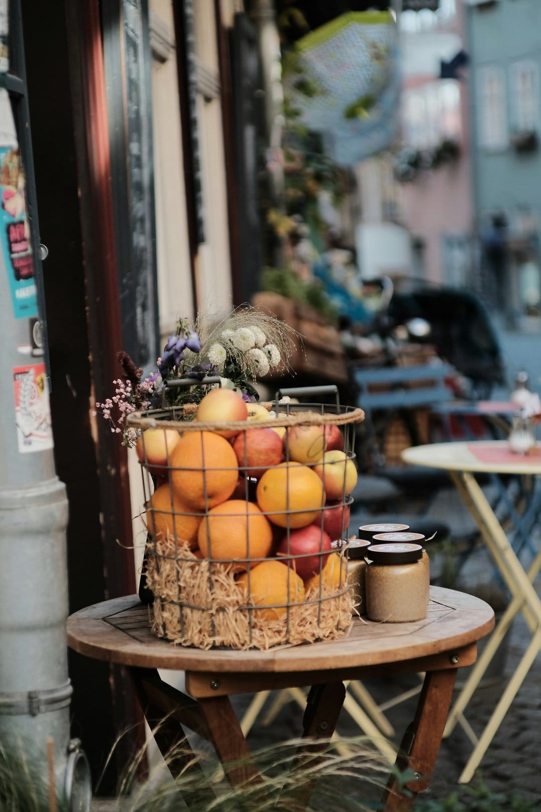 Oranges and apples in a wire basket next to dried flowers on a wooden table outside a European cafe.