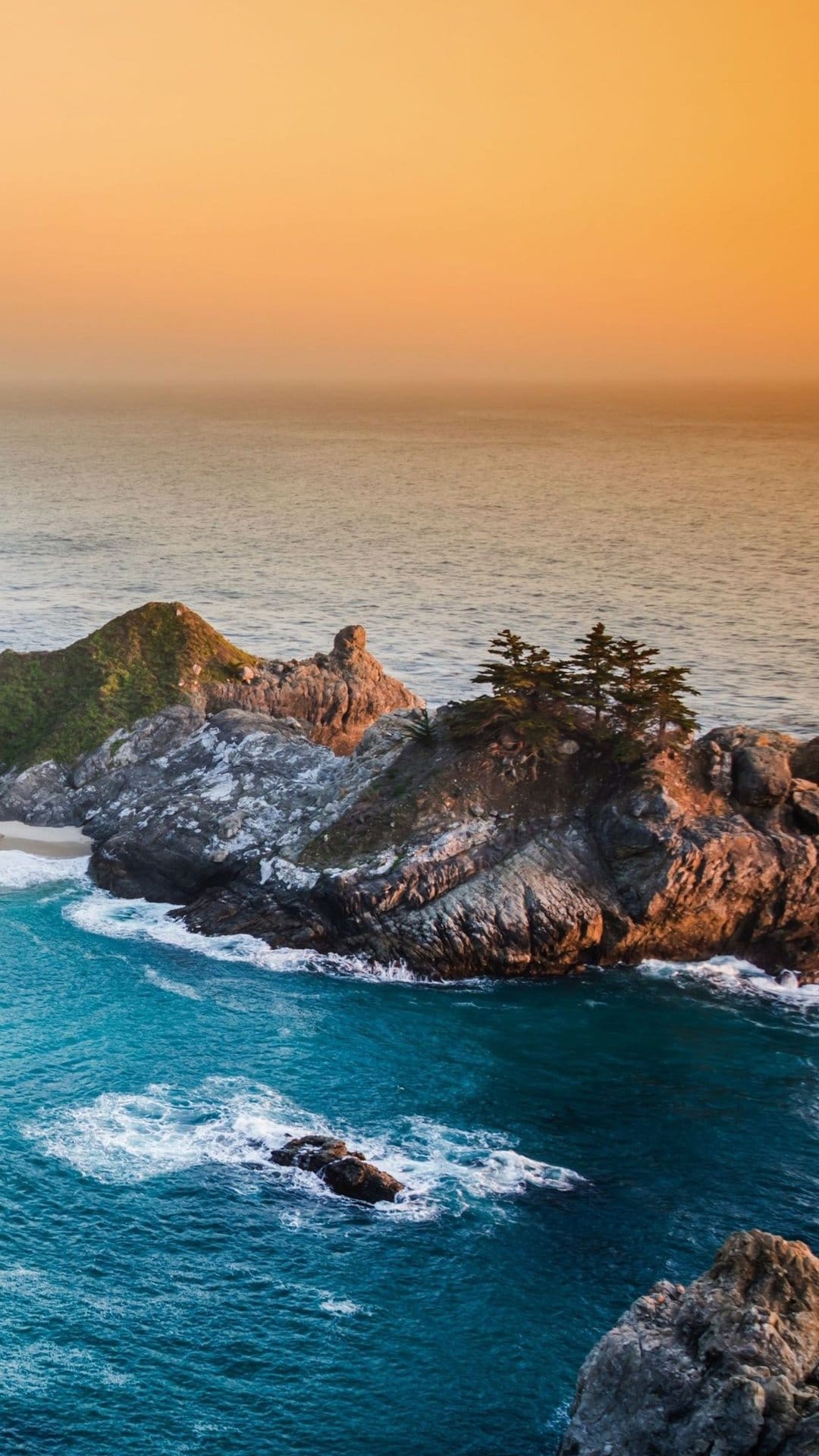 Jagged cliffs overlook a turquoise cove with gentle waves under a warm golden hour sky on the California coast.