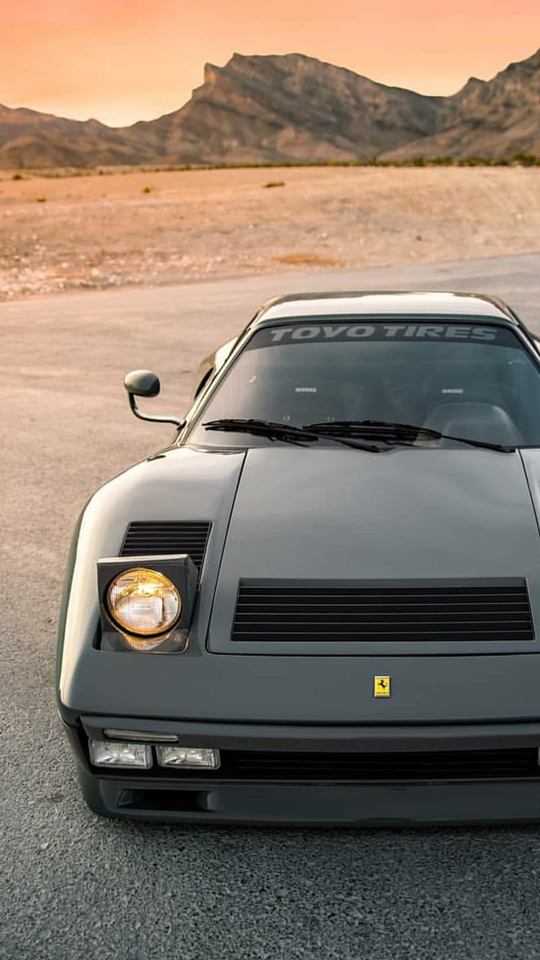 Grey Ferrari with pop-up headlights on a gravel desert road against a glowing sunset mountain backdrop.