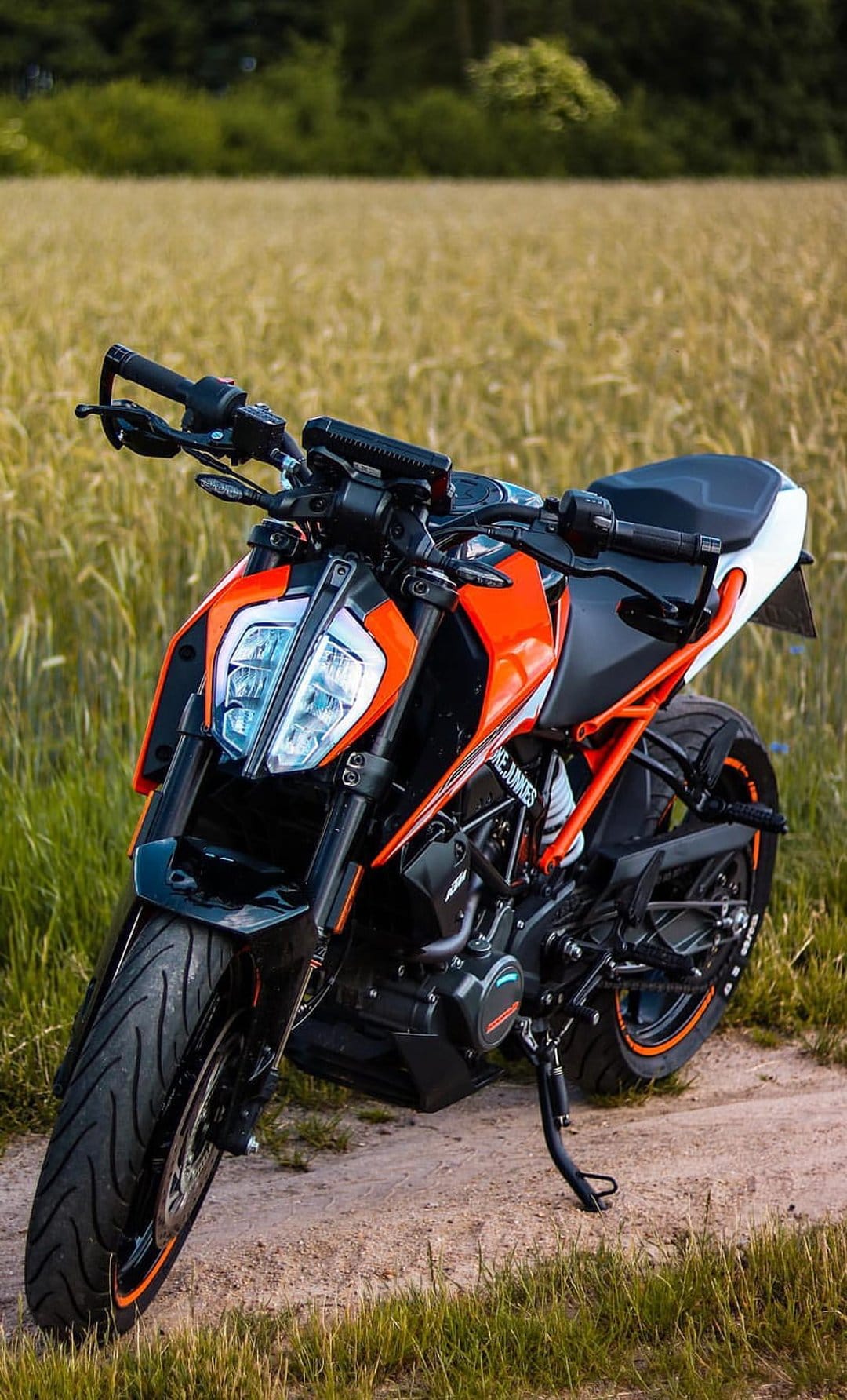 Orange KTM motorcycle with a black seat parked on a dirt trail between golden grass and green fields under daylight.