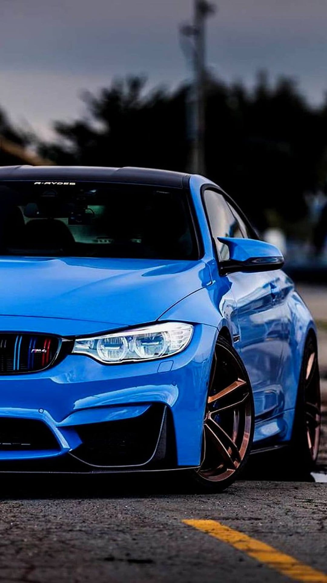 BMW M4 Coupe in metallic blue features bronze multi-spoke wheels and glowing LED headlights in a low-angle shot.