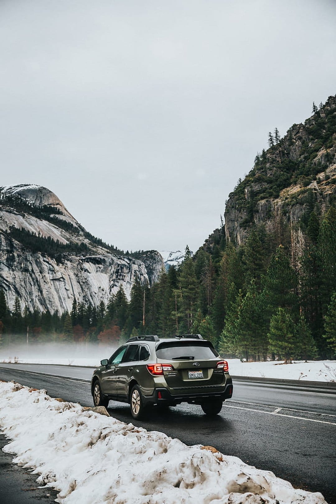 Green Subaru Outback on a snowy Yosemite road with towering granite cliffs and pine trees under a gray sky.