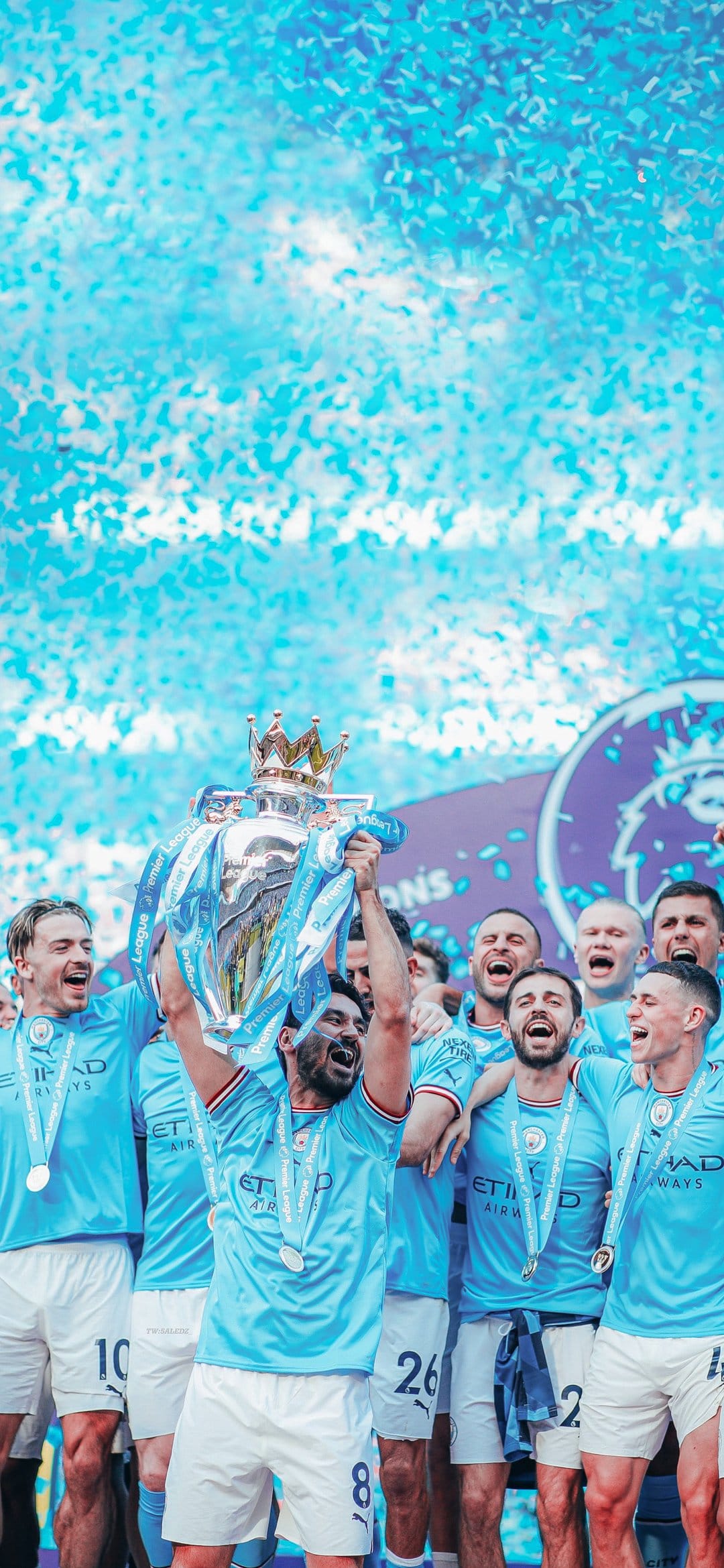 Manchester City football players lifting the Premier League trophy surrounded by blue and white confetti in a stadium.