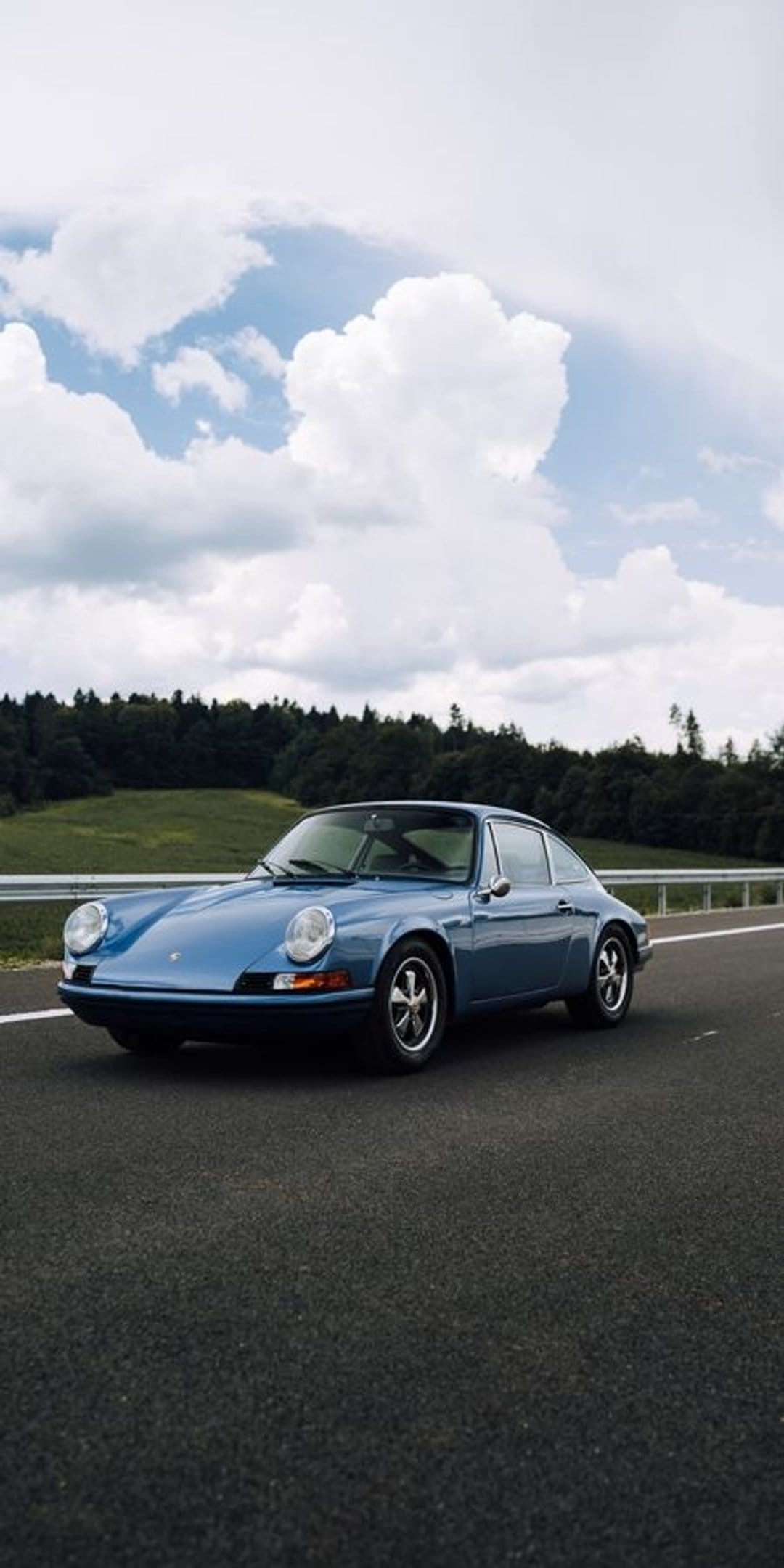 Blue vintage Porsche 911 coupe driving on a paved road between green fields and a dense forest under clouds.