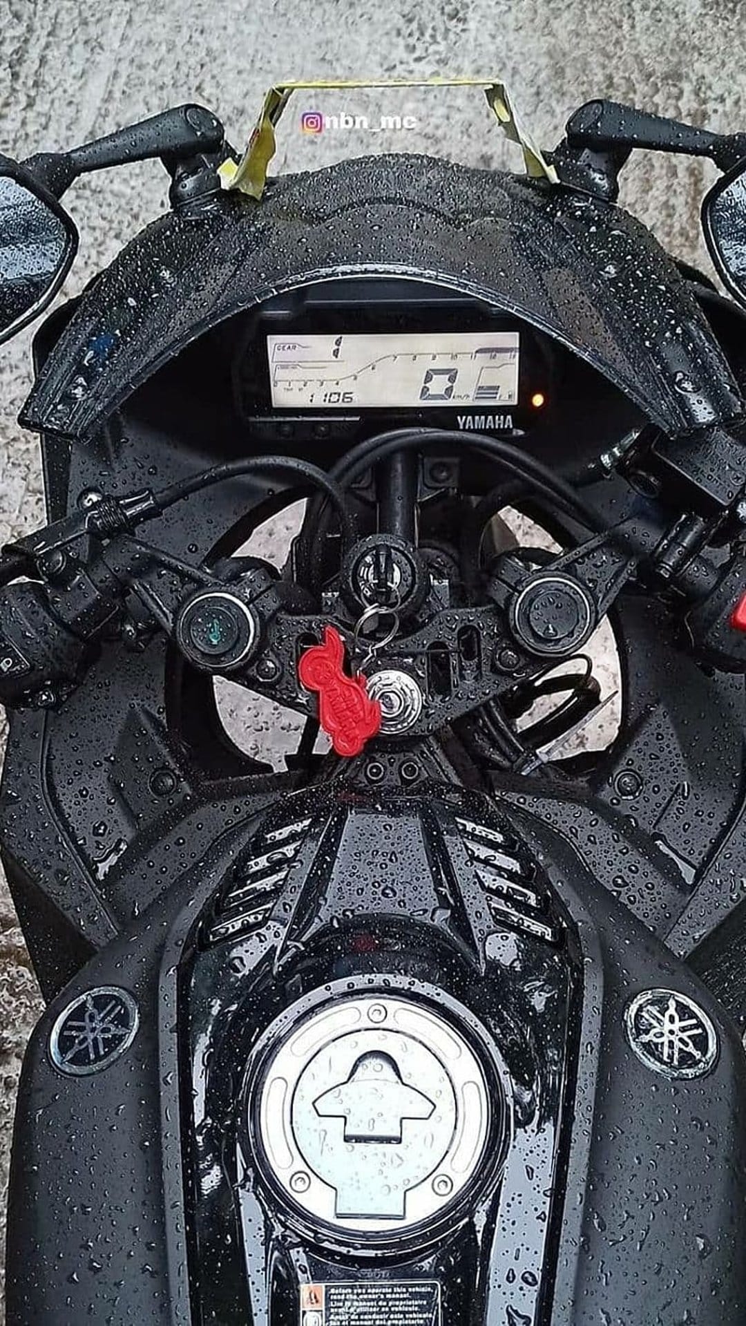 Yamaha R15 V3 handlebars and fuel tank covered in raindrops with a red cat keychain and digital speedometer.