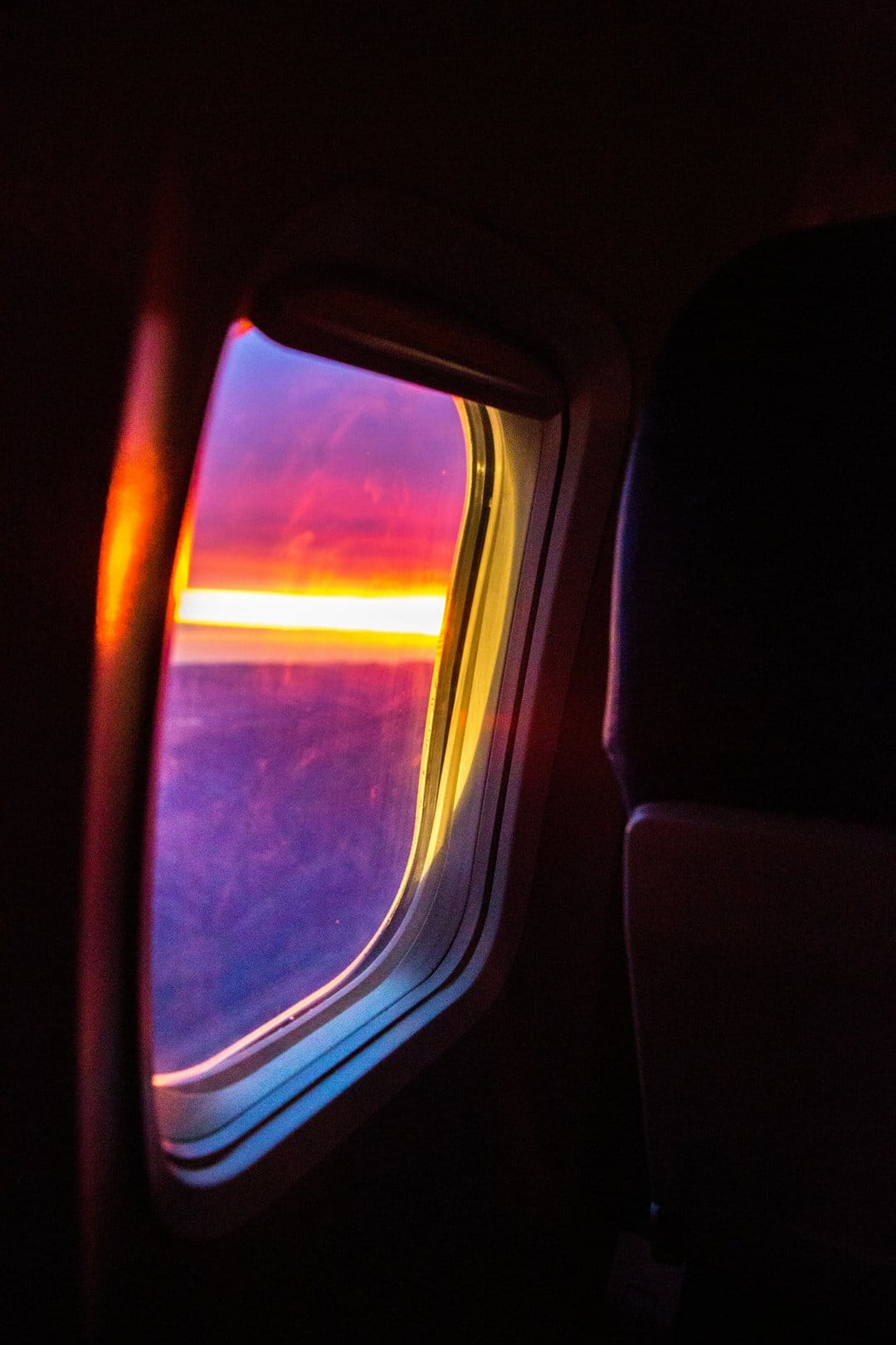 Airplane window framing a purple and orange sunset over the ocean horizon with soft light reflecting on the rim.