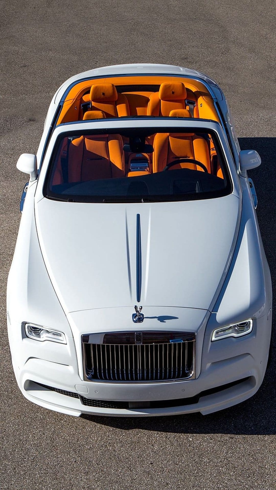 White Rolls-Royce convertible with vibrant orange leather seats viewed from an aerial angle on dark asphalt.