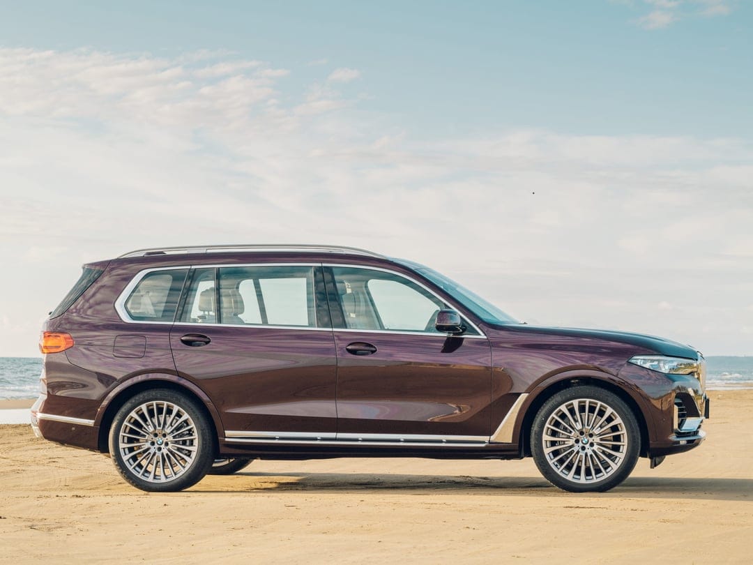 Side profile of a plum BMW X7 parked on a sandy beach under a light blue sky with soft clouds and ocean.