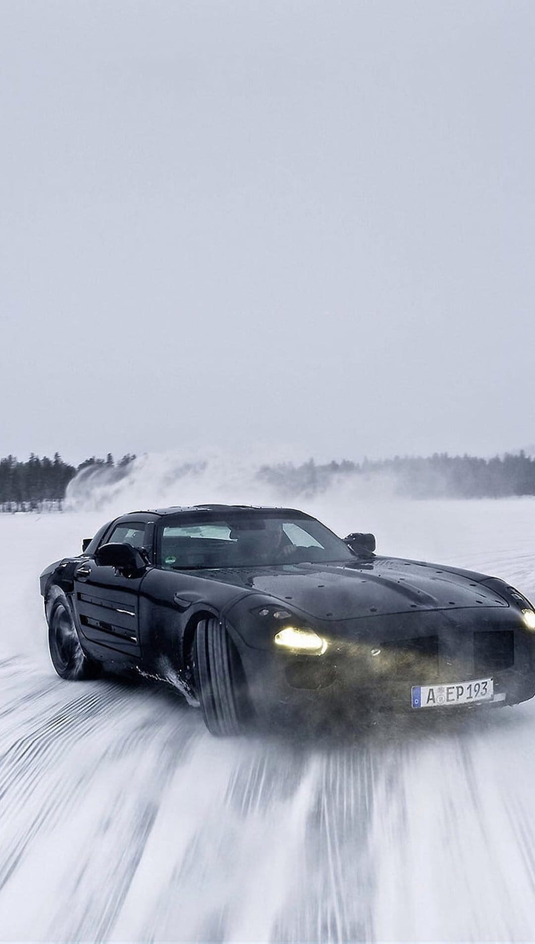 Mercedes SLS AMG drifts on a snow-covered road with headlights glowing through a hazy winter atmosphere.