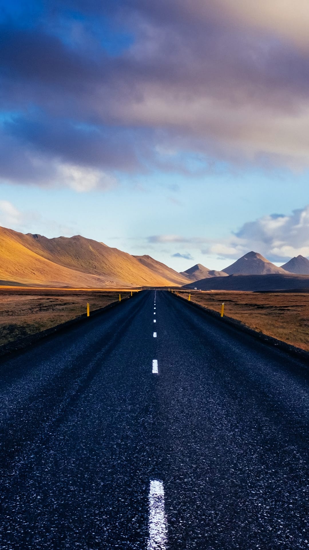 Long straight road with white markings stretching toward golden mountains under a cloudy sky in rural Iceland.