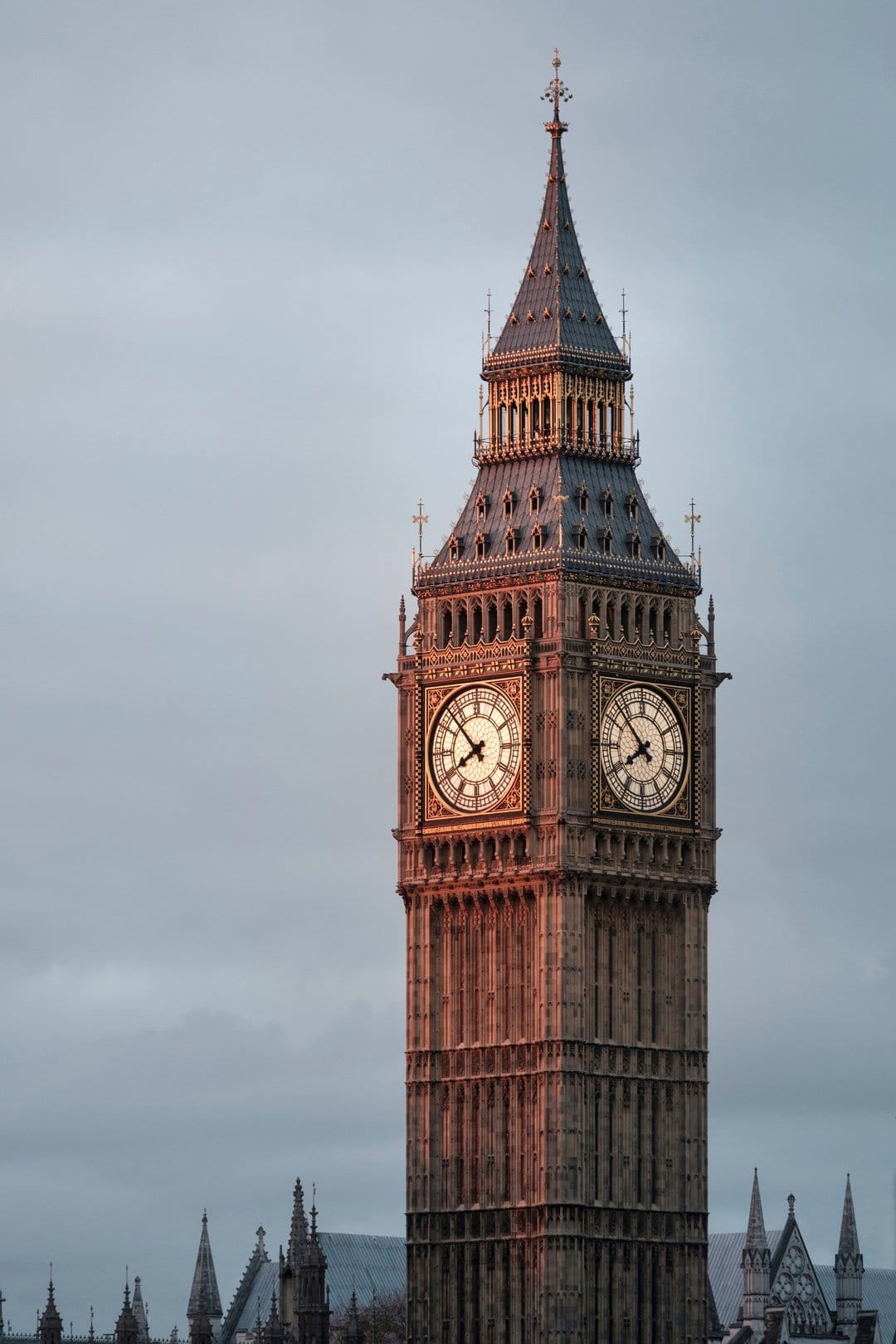 Elizabeth Tower clock face glows against a cloudy sunset sky with the Palace of Westminster in the foreground.