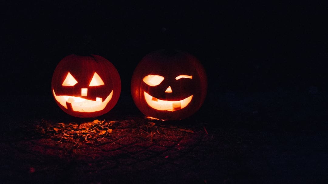 Two carved jack-o'-lanterns with glowing faces sit on dark ground, featuring a winking eye and a wide smile.