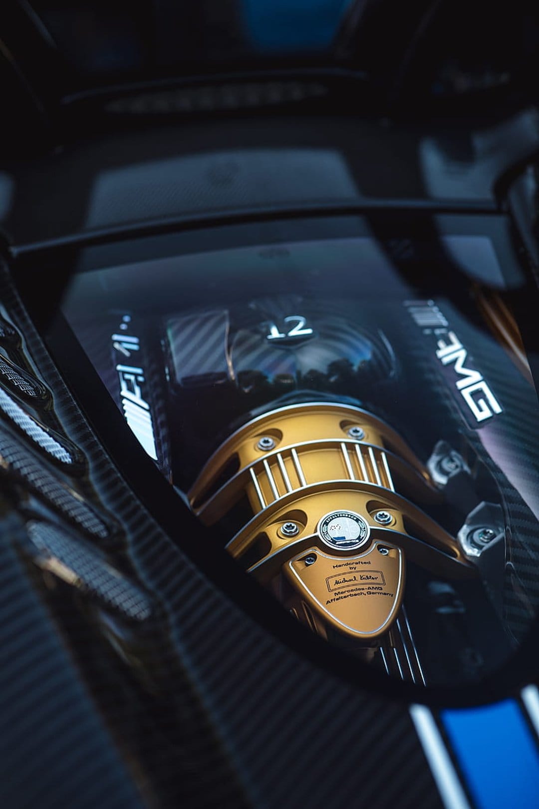 Mercedes-AMG V12 engine showing carbon fiber weaving, a gold cover, and a silver handcrafted signature plaque.