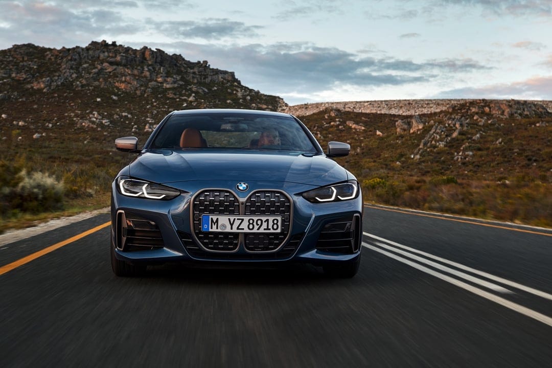 Blue BMW 4 Series coupe driving on a winding asphalt road through rocky hills during a golden sunset.