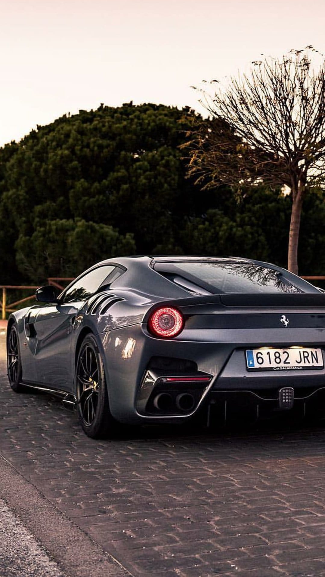 Grey Ferrari F12 TDF with lit taillights parked on a textured cobblestone street surrounded by green trees.