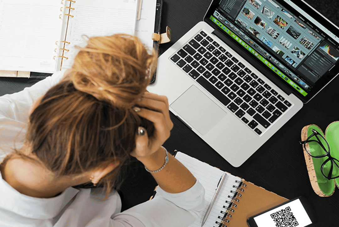 Woman leans over a messy desk with a glowing laptop and scattered notebooks in a vertical overhead view.