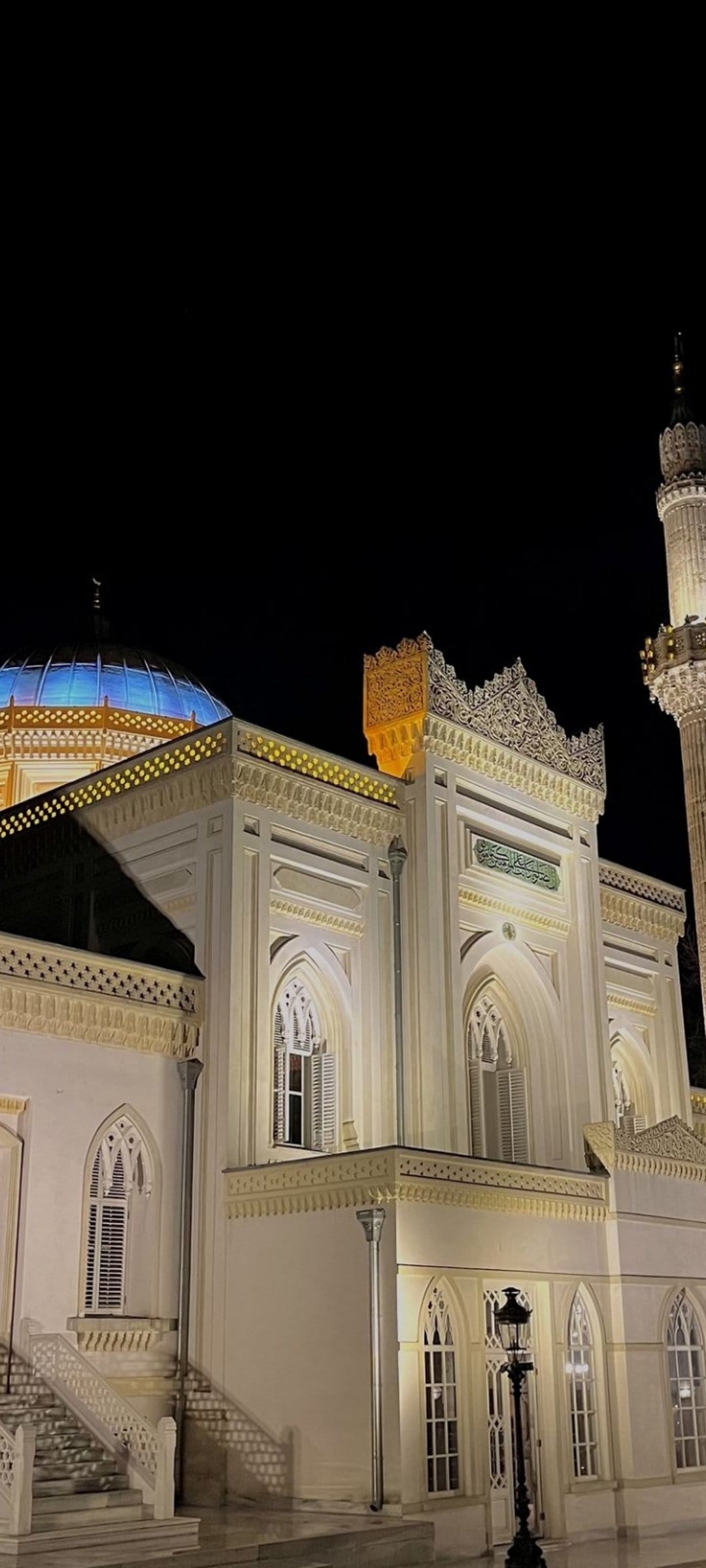 Luminous blue mosque dome and tall minaret illuminated by warm lights against a dark night sky in vertical view.