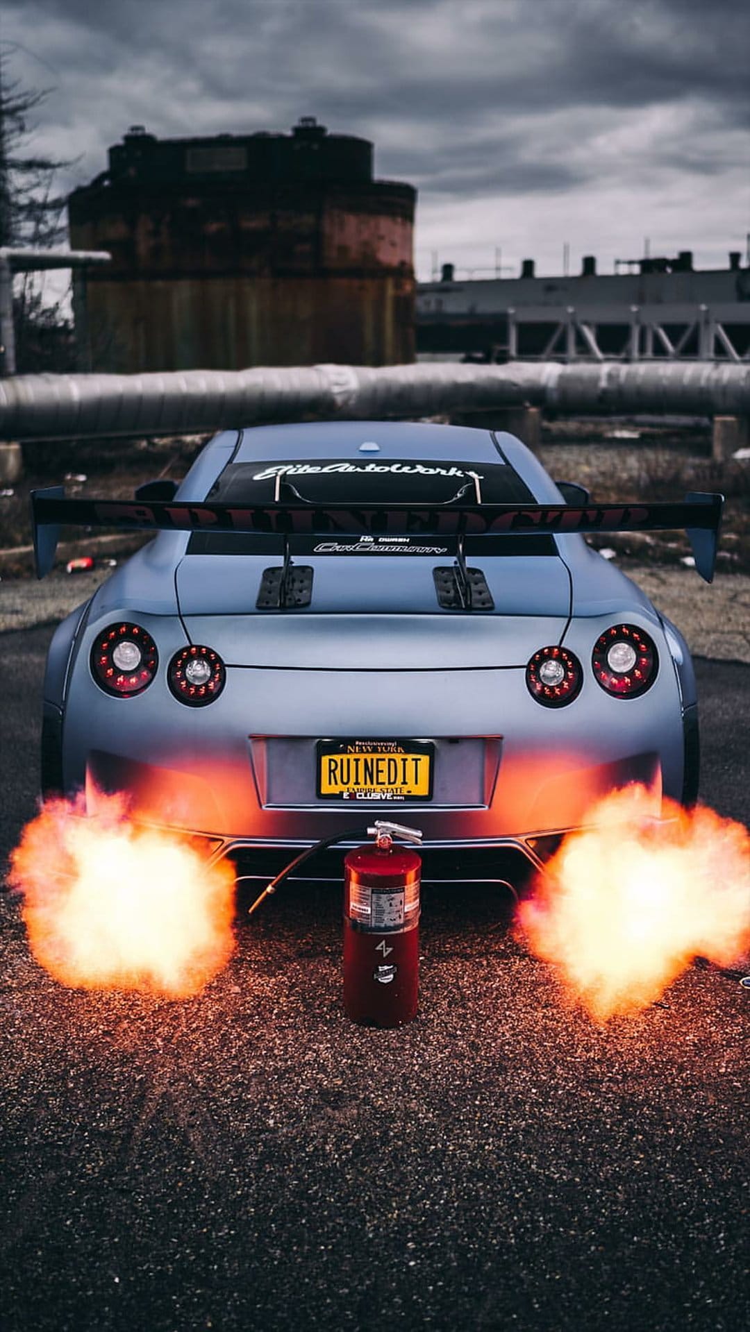 Nissan GT-R with a Ruined It plate shoots orange exhaust flames near a red fire extinguisher on asphalt.