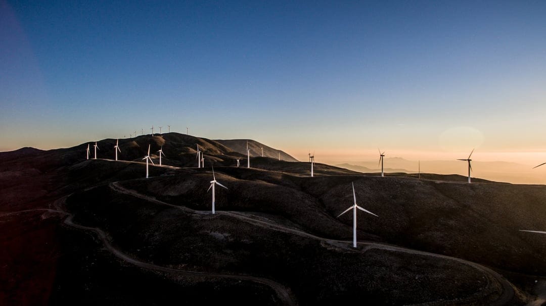Wind turbine silhouettes stand on dark rolling hills against a vibrant orange and purple sunset sky at twilight.