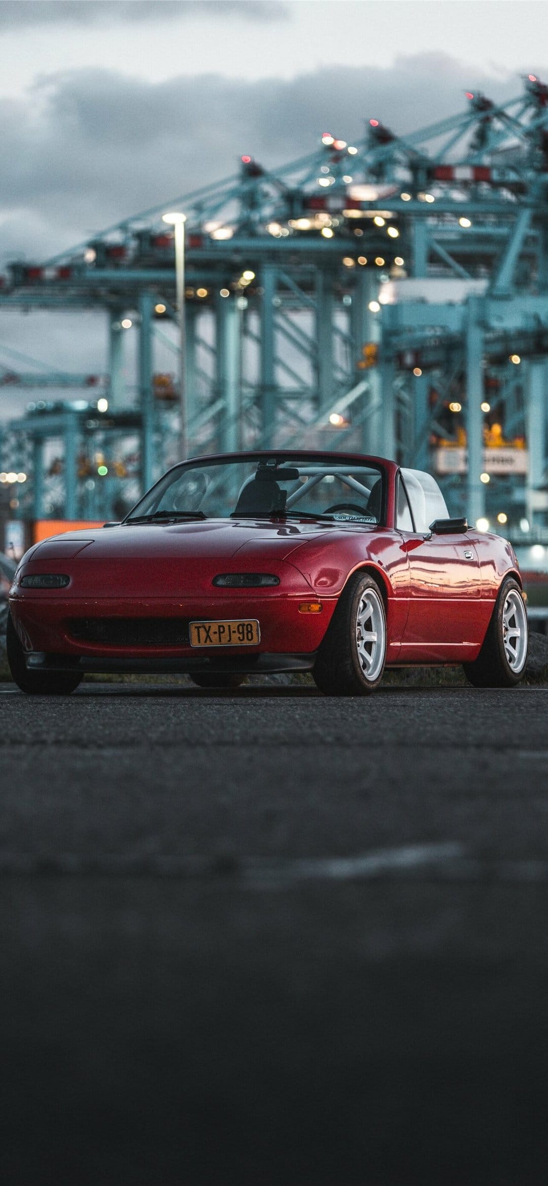 Red Mazda Miata NA with white wheels parked in front of glowing industrial port cranes at twilight.