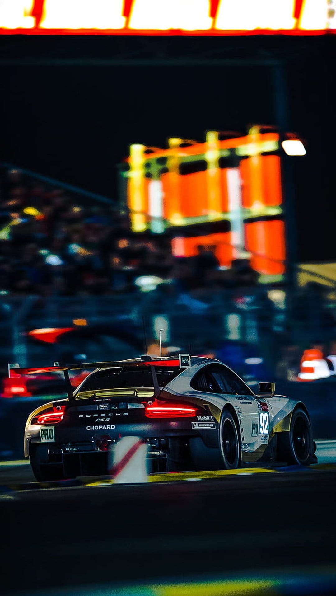 Porsche 911 RSR race car with glowing red taillights cornering at night past blurred crowds and track lights.