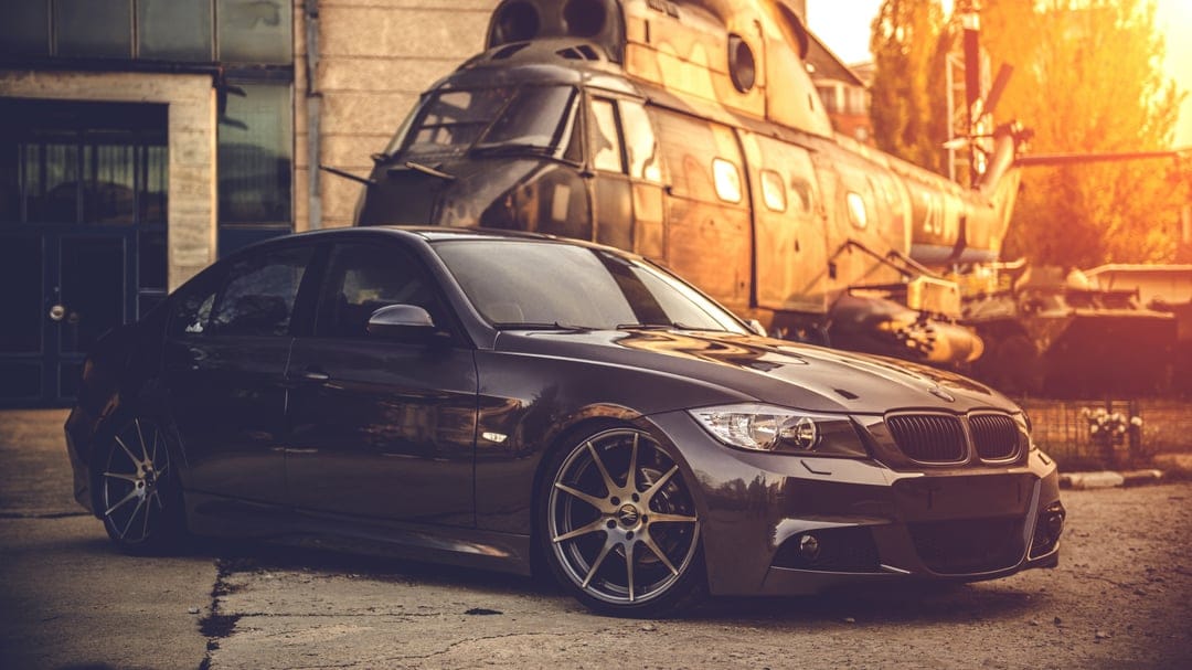 Dark sedan with custom rims parked next to a weathered vintage helicopter during a warm golden hour sunset.