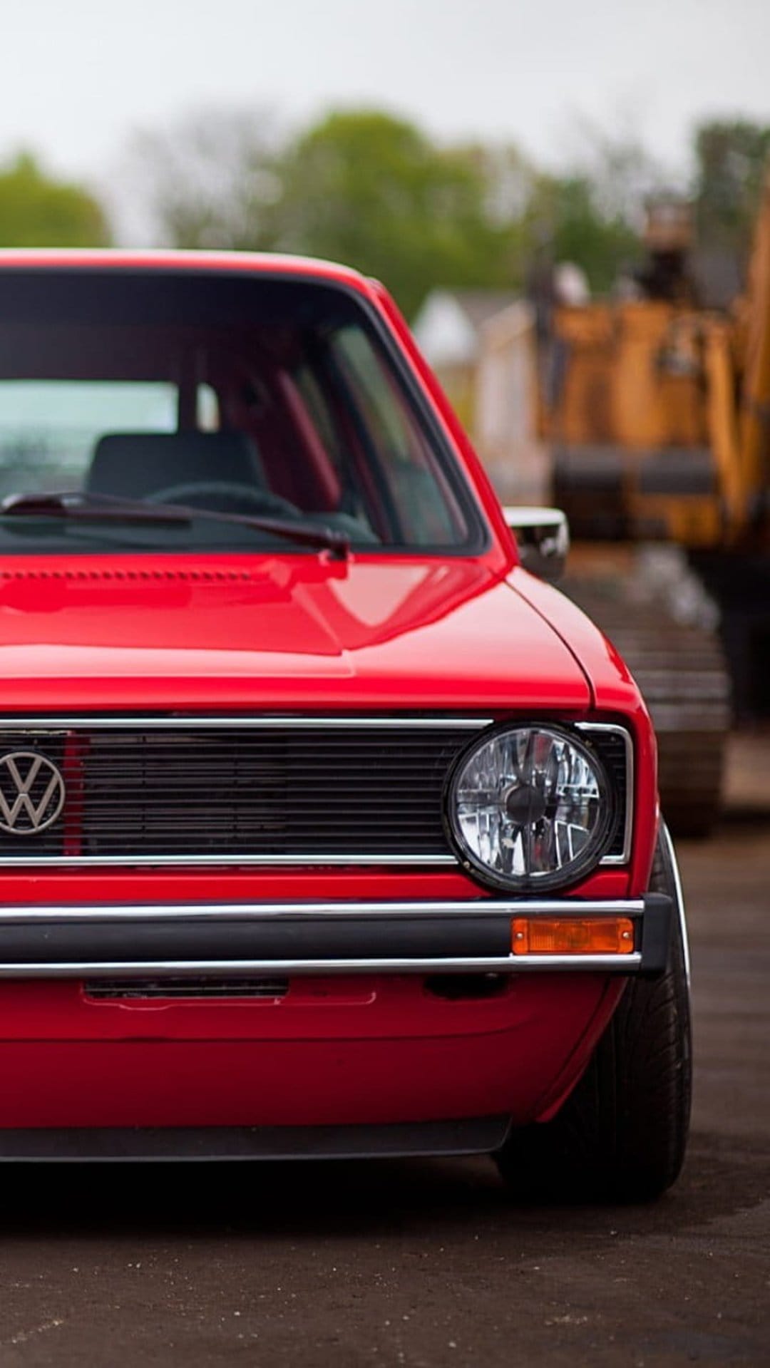 Red Volkswagen Golf MK1 front end featuring a round headlight and VW badge against a blurred green background.