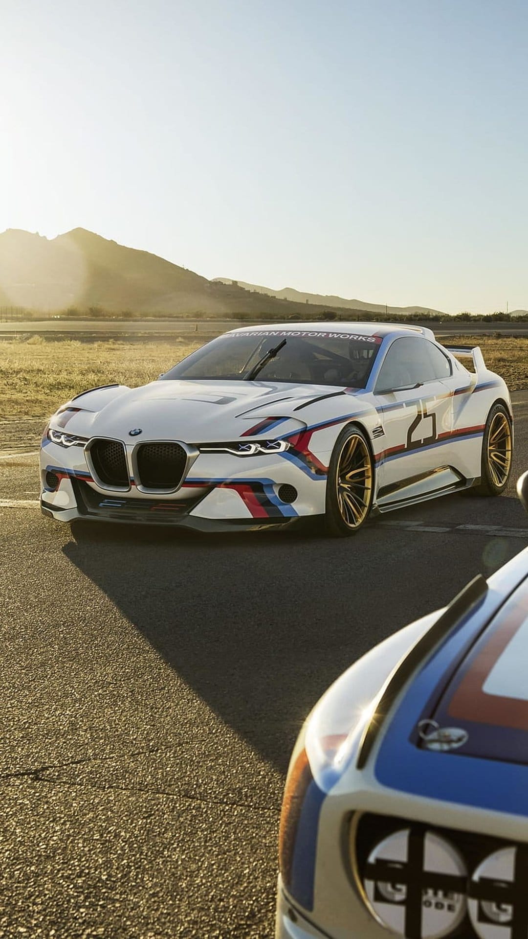 BMW 3.0 CSL Hommage R race car with number 25 and gold wheels on an asphalt track against a mountain backdrop.
