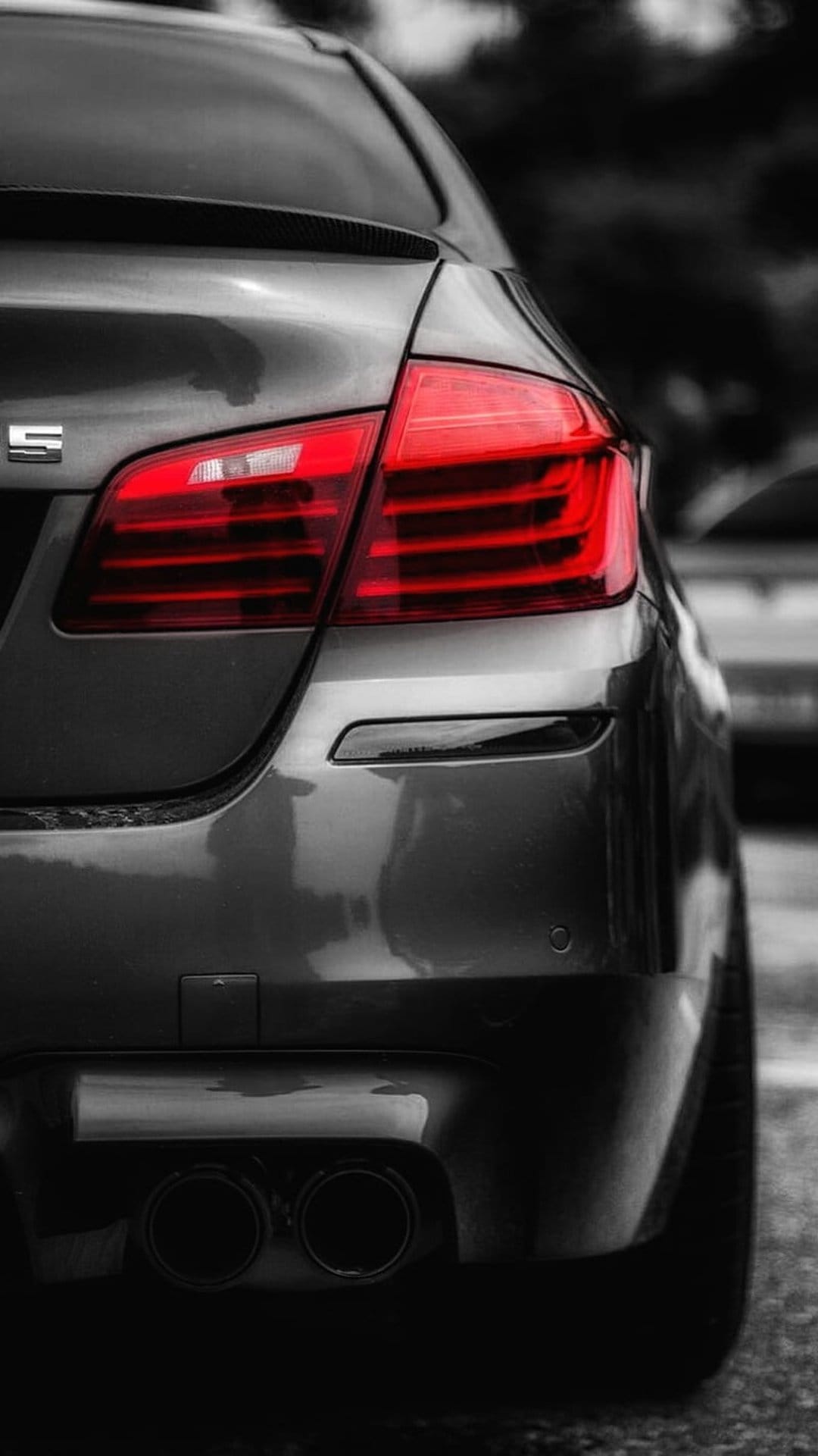 BMW M Series red LED tail light and dual exhaust pipes on a dark metallic car body with selective coloring.