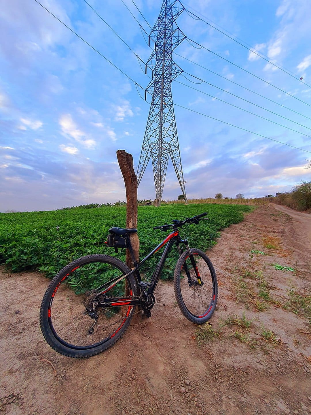 Red and black mountain bike leaning on a post next to a tall electrical transmission tower in a grassy field.