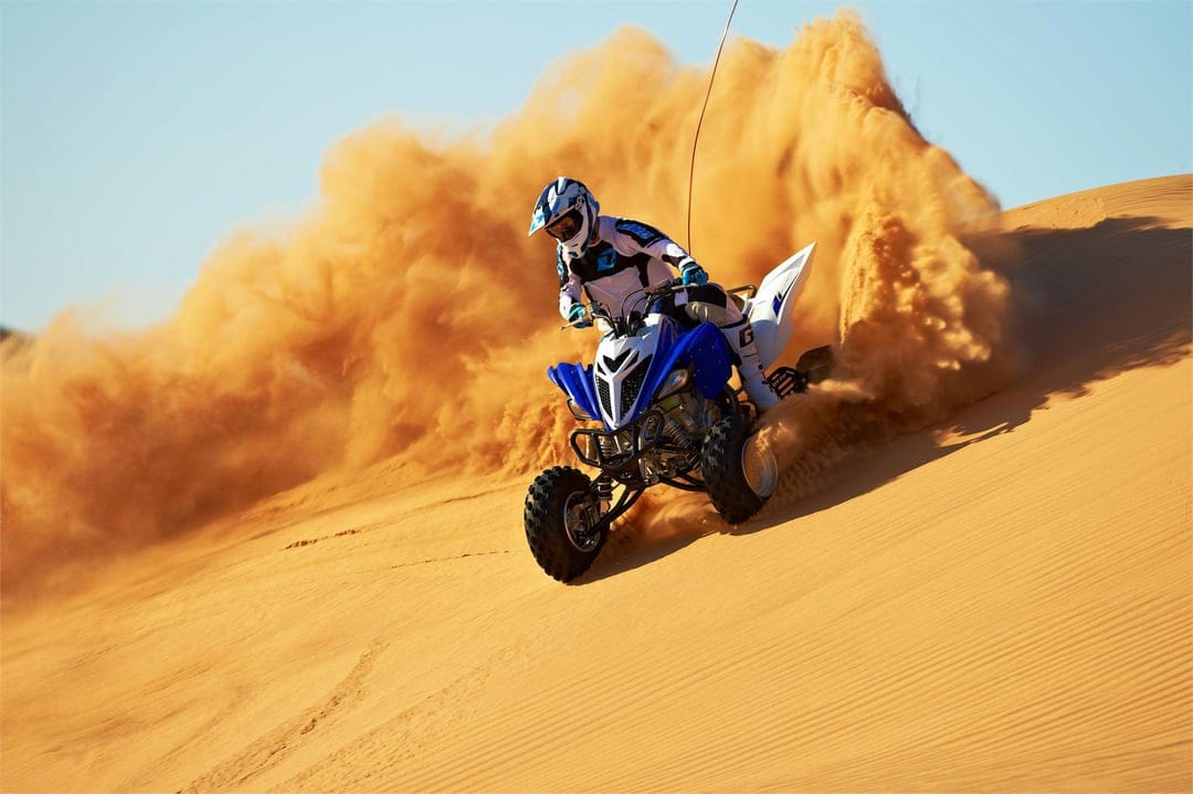 Blue quad bike rider drifting on a golden sand dune, creating a massive orange dust cloud under a clear sky.