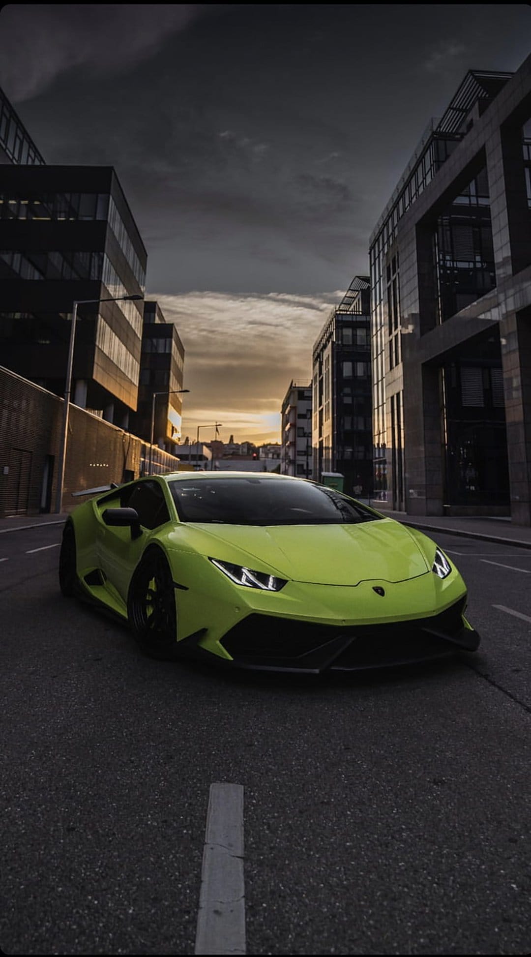 Lime green Lamborghini Huracan drives on a city street under a dramatic orange and purple sunset sky.