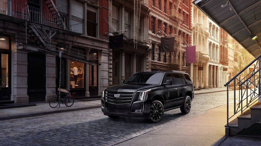 Black Cadillac Escalade SUV parked on a city cobblestone street with brick buildings and fire escapes in sunlight.