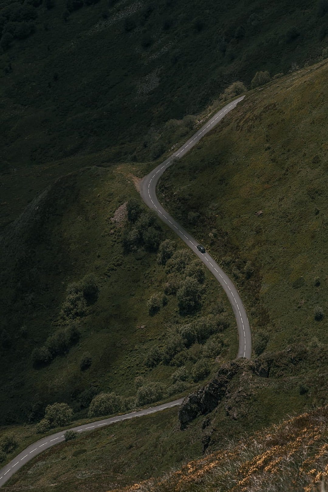 Winding asphalt road curves through steep green hills with a single white car driving along the mountain ridge.
