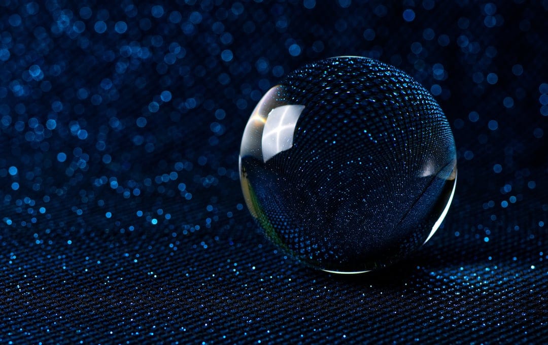 Clear crystal ball reflecting light while resting on a dark blue textile covered in sparkling silver glitter.