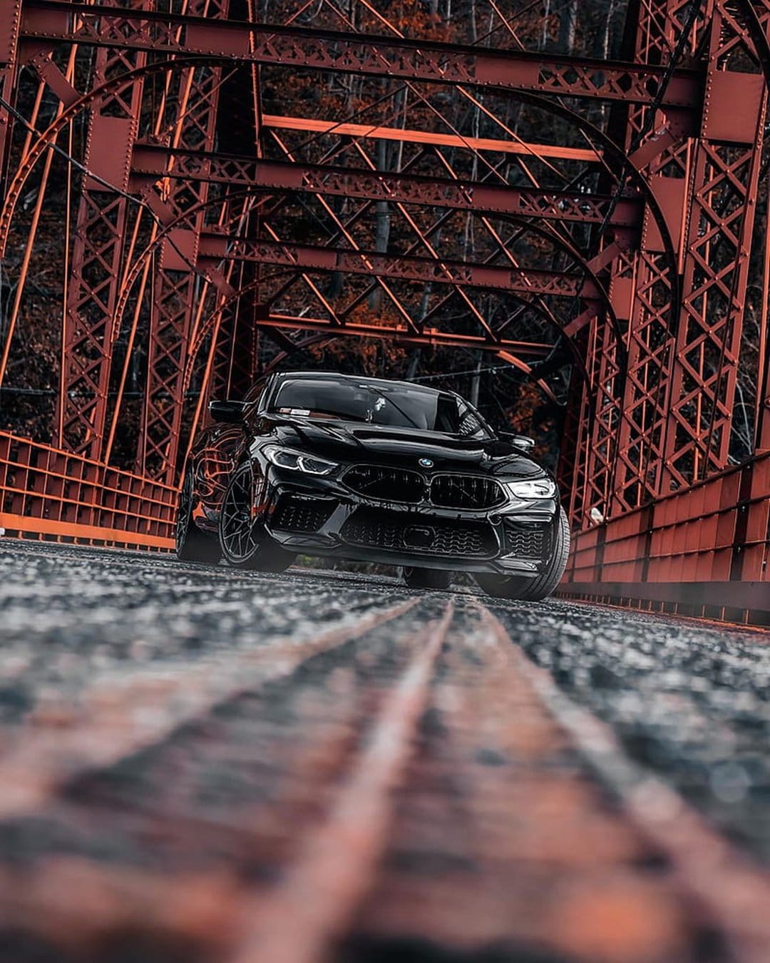 Black BMW M8 sports car parked on a red steel truss bridge with a low-angle view and weathered road texture.