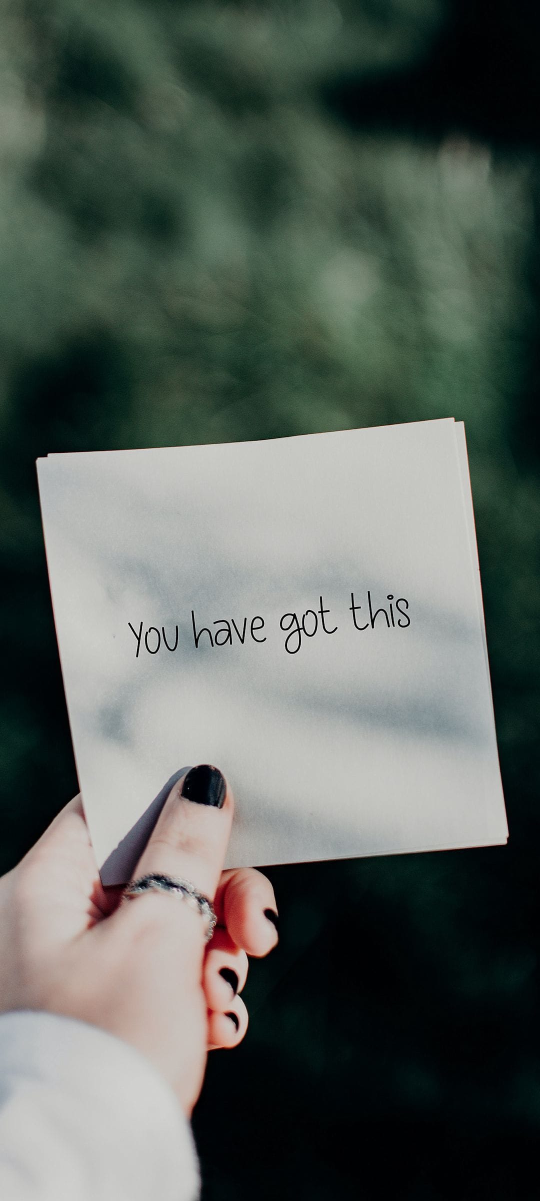 Hand with black nail polish holds a You Have Got This note over blurred dark green leaves in a forest setting.