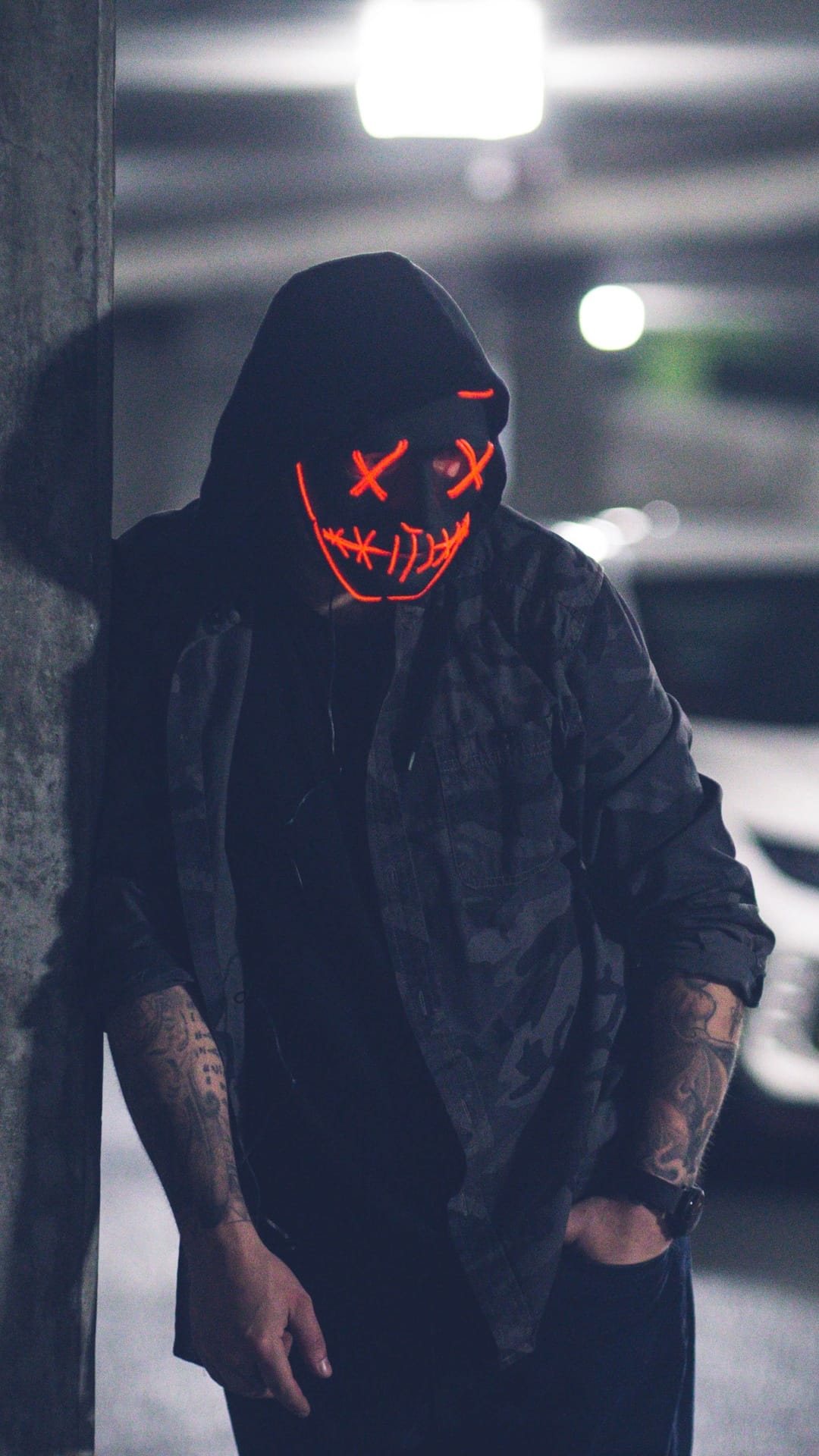 Person in a red glowing X-eye mask and dark hoodie leans against a concrete pillar in a dimly lit garage.