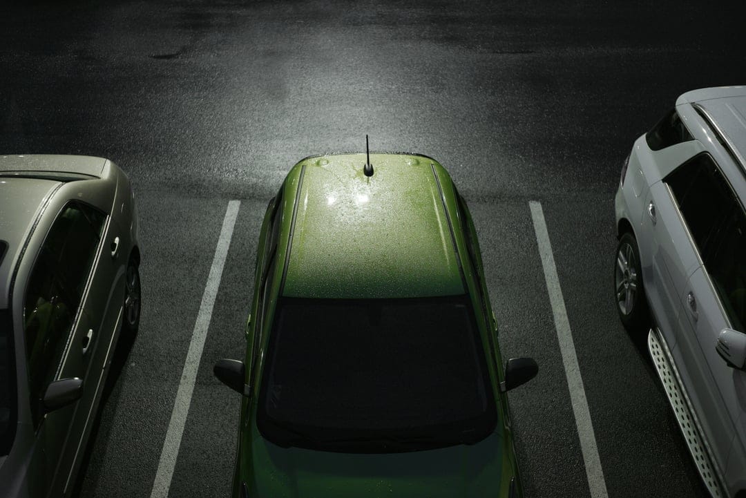 Green, silver, and white cars parked on dark wet asphalt viewed from an overhead aerial perspective at night.