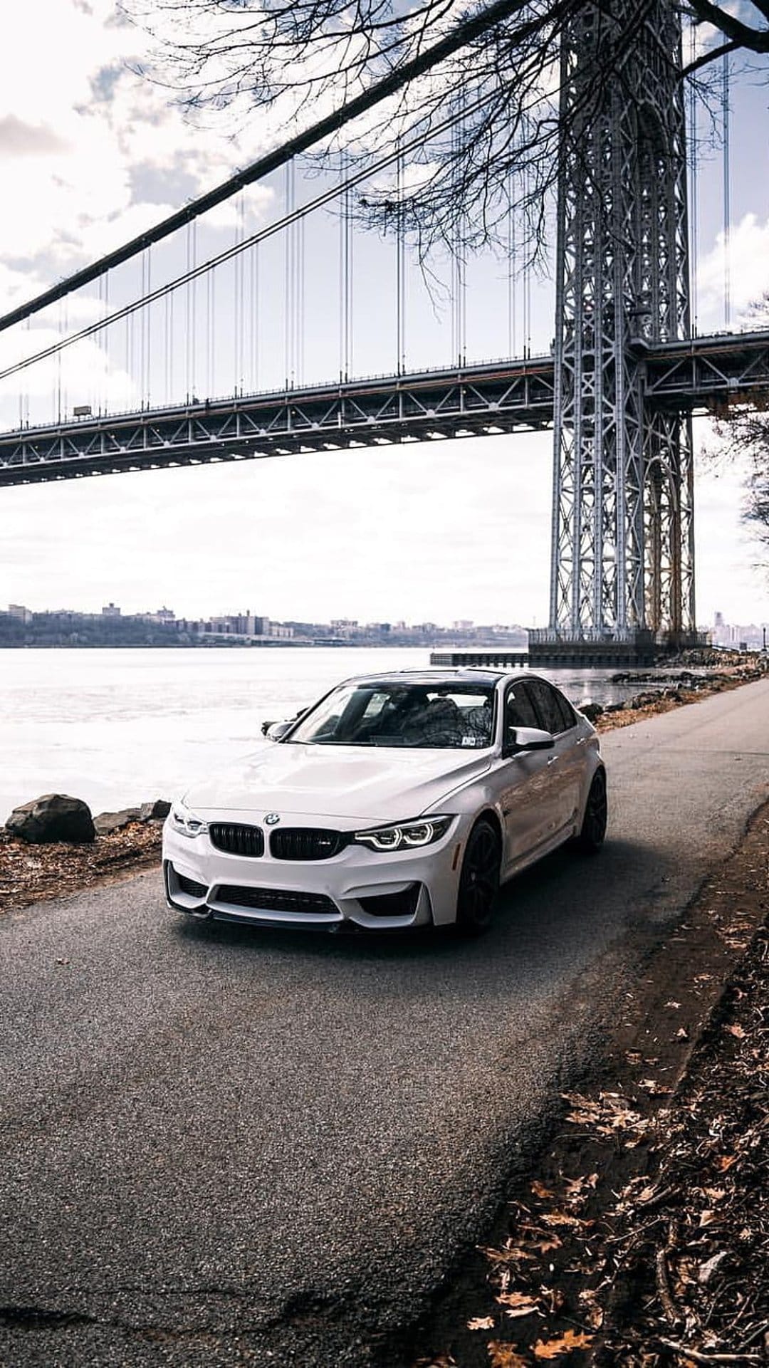 White BMW M3 sedan parked on an asphalt road beside a river with a large suspension bridge in the background.