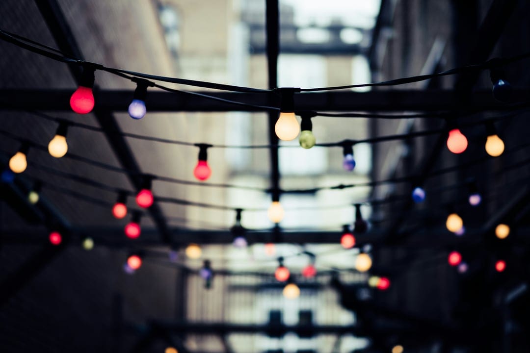 Glowing glass bulbs hang on a string above a darkened alleyway with soft colorful bokeh circles in the background.