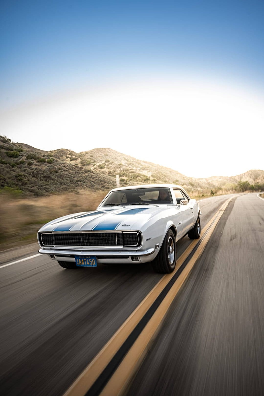 White classic Camaro with blue racing stripes speeds down a winding highway during a warm golden sunset.
