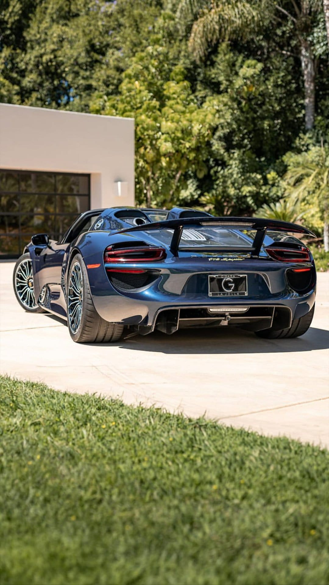 Porsche 918 Spyder hypercar in dark blue parked on a concrete driveway with trees and a white building behind.