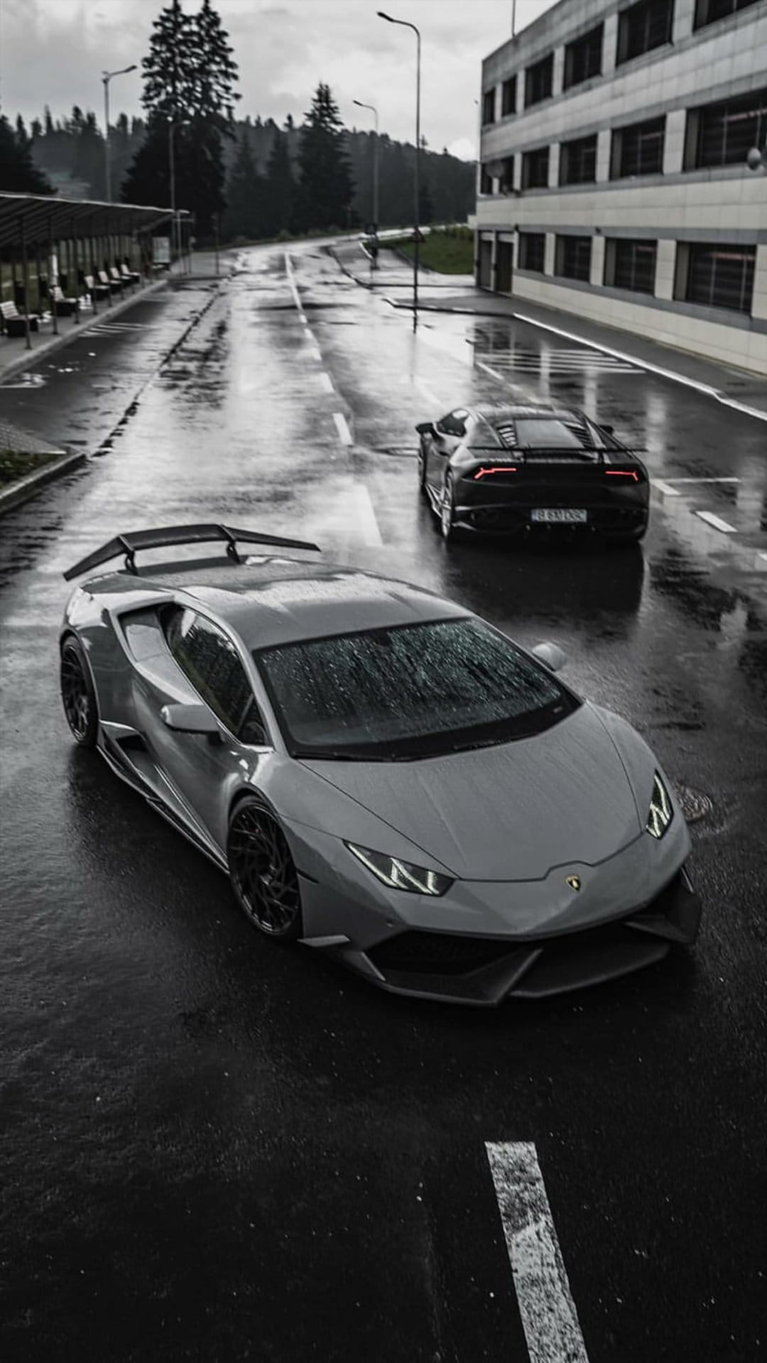 Grey and black Lamborghinis driving on wet asphalt with reflections and modern buildings in the background.