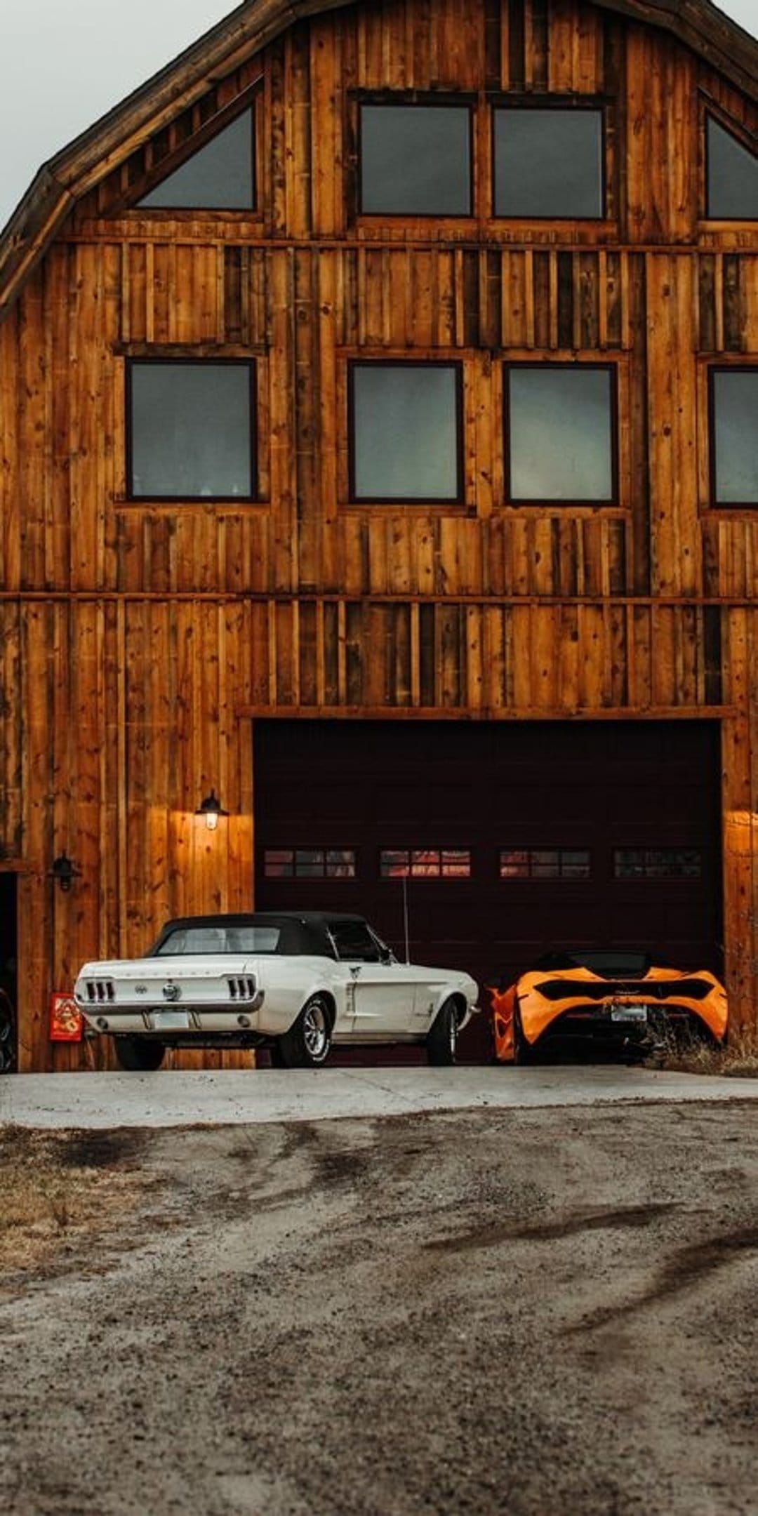 White Ford Mustang and orange McLaren parked in front of a weathered wooden barn with large glass windows.